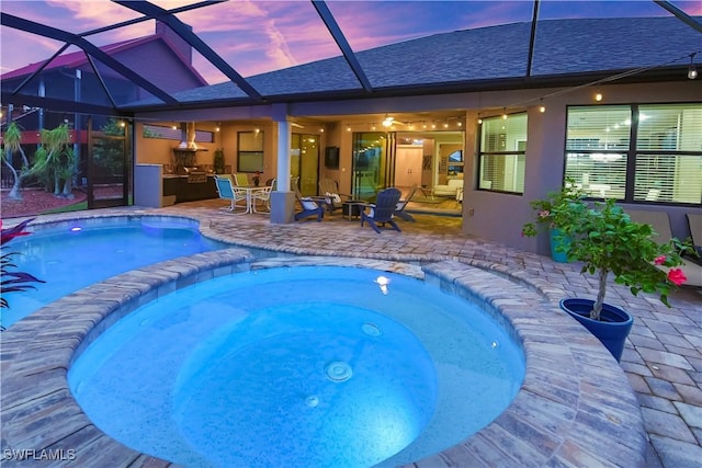 pool at dusk featuring an outdoor kitchen, glass enclosure, a patio, and an in ground hot tub