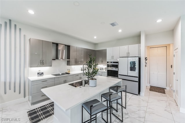 kitchen with gray cabinets, a center island with sink, appliances with stainless steel finishes, a kitchen breakfast bar, and wall chimney exhaust hood