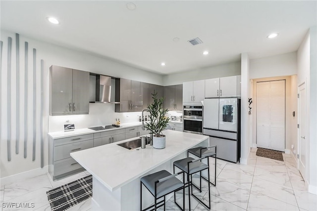 kitchen with a breakfast bar area, gray cabinetry, appliances with stainless steel finishes, a kitchen island with sink, and wall chimney range hood