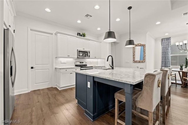 kitchen featuring appliances with stainless steel finishes, white cabinets, decorative light fixtures, an island with sink, and light stone counters