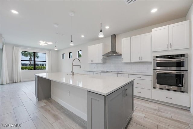 kitchen featuring wall chimney exhaust hood, stainless steel double oven, a center island with sink, and white cabinets