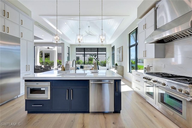 kitchen featuring white cabinetry, high quality appliances, ventilation hood, a center island with sink, and decorative light fixtures