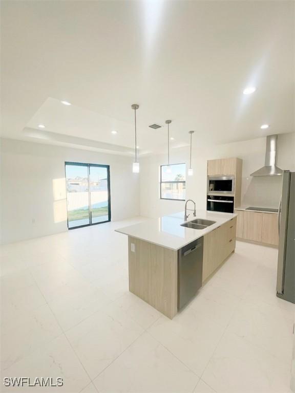 kitchen with a center island with sink, appliances with stainless steel finishes, light brown cabinetry, wall chimney range hood, and sink