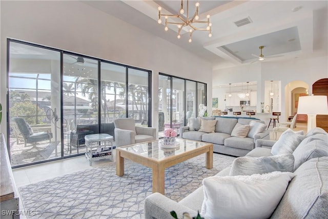 living room with visible vents, arched walkways, a towering ceiling, a tray ceiling, and ceiling fan with notable chandelier