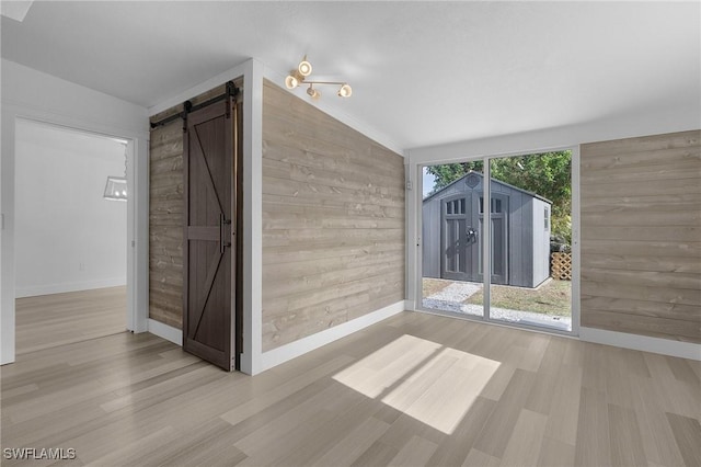 empty room with a barn door, wooden walls, baseboards, vaulted ceiling, and light wood-type flooring