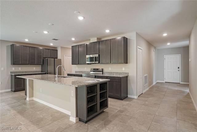 kitchen with appliances with stainless steel finishes, a kitchen island with sink, light stone countertops, dark brown cabinetry, and sink