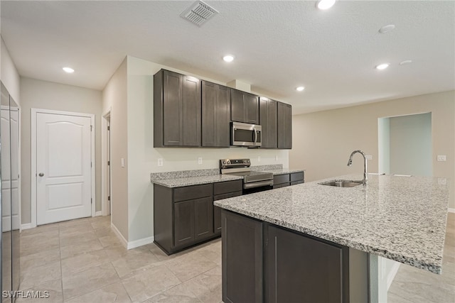 kitchen featuring a center island with sink, sink, dark brown cabinetry, stainless steel appliances, and light stone counters