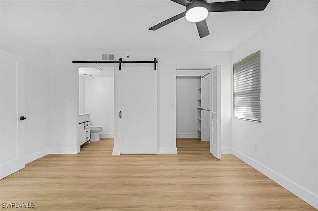unfurnished bedroom featuring a barn door, a walk in closet, connected bathroom, baseboards, and light wood-type flooring