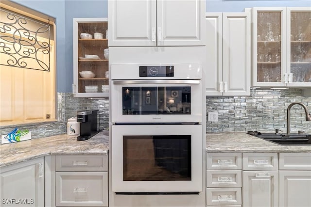kitchen with glass insert cabinets, a sink, white double oven, decorative backsplash, and white cabinets