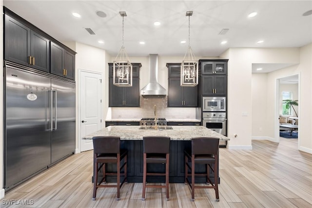 kitchen with a center island with sink, glass insert cabinets, decorative light fixtures, built in appliances, and wall chimney range hood