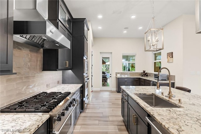 kitchen featuring dark cabinets, a sink, wall chimney range hood, decorative backsplash, and pendant lighting