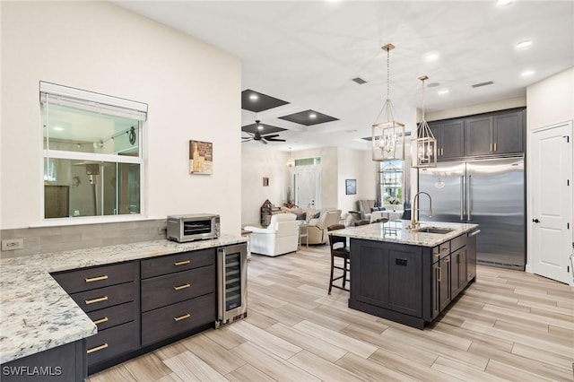 kitchen featuring a center island with sink, wine cooler, hanging light fixtures, built in refrigerator, and a sink