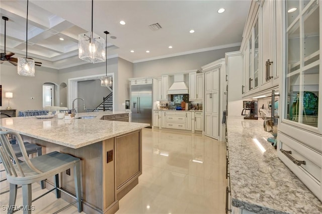 kitchen featuring built in fridge, light stone countertops, a large island with sink, decorative light fixtures, and a breakfast bar area