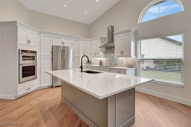 kitchen featuring a center island with sink, appliances with stainless steel finishes, wall chimney range hood, white cabinets, and sink