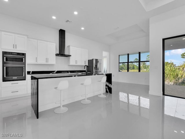 kitchen featuring white cabinets, wall chimney exhaust hood, a kitchen bar, stainless steel refrigerator with ice dispenser, and an island with sink