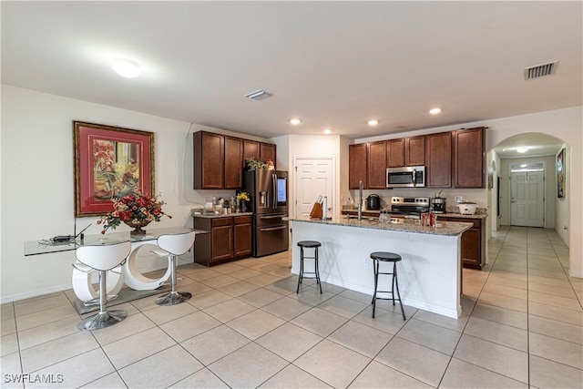 kitchen with a center island with sink, a breakfast bar, stainless steel appliances, light tile patterned floors, and dark stone counters