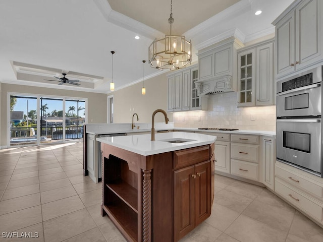 kitchen with sink, decorative light fixtures, a raised ceiling, stainless steel double oven, and a kitchen island with sink