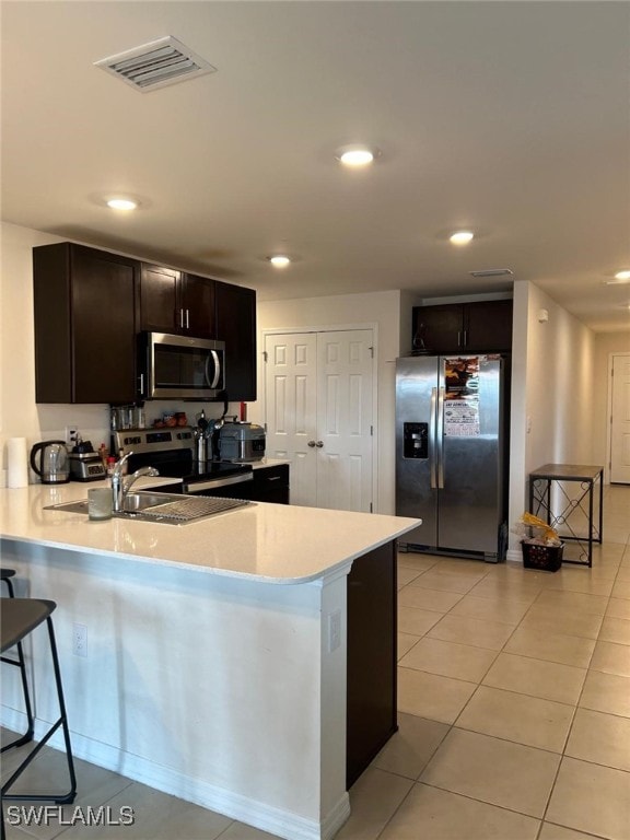 kitchen with kitchen peninsula, a kitchen bar, light tile patterned flooring, dark brown cabinetry, and appliances with stainless steel finishes