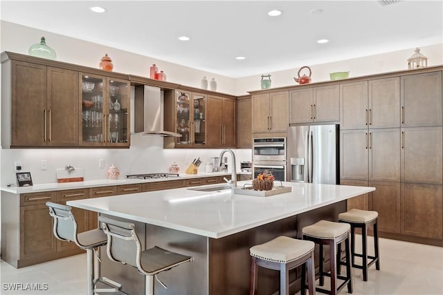 kitchen featuring wall chimney range hood, sink, a breakfast bar area, appliances with stainless steel finishes, and a kitchen island with sink