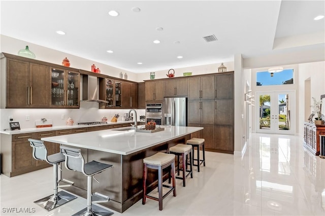 kitchen featuring wall chimney range hood, sink, appliances with stainless steel finishes, a kitchen breakfast bar, and an island with sink