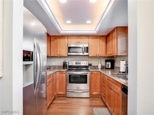 kitchen with a raised ceiling, decorative backsplash, light wood-type flooring, light stone countertops, and stainless steel appliances