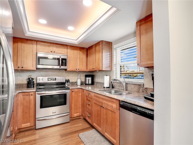 kitchen with appliances with stainless steel finishes, sink, light stone counters, a tray ceiling, and light hardwood / wood-style flooring