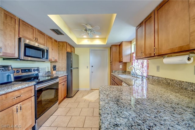 kitchen with sink, ceiling fan, appliances with stainless steel finishes, light stone counters, and a tray ceiling