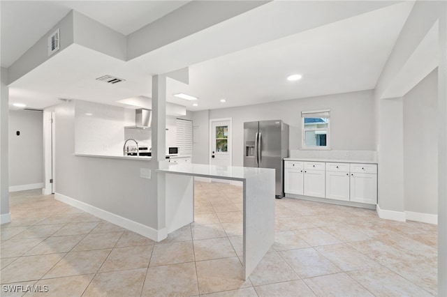 kitchen featuring light tile patterned floors, stainless steel fridge, kitchen peninsula, white cabinets, and wall chimney range hood