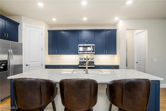 kitchen with blue cabinetry, appliances with stainless steel finishes, wood-type flooring, and a kitchen island with sink