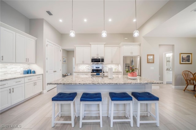 kitchen featuring hanging light fixtures, an island with sink, stainless steel appliances, white cabinets, and light stone counters