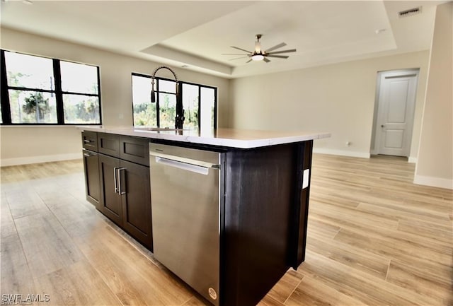 kitchen with light wood-type flooring, stainless steel dishwasher, a tray ceiling, and a kitchen island with sink