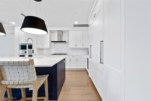 kitchen featuring a breakfast bar, pendant lighting, an island with sink, white cabinets, and wall chimney range hood