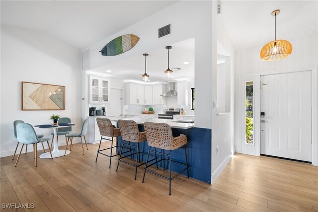 kitchen with hanging light fixtures, white cabinetry, wall chimney range hood, and kitchen peninsula