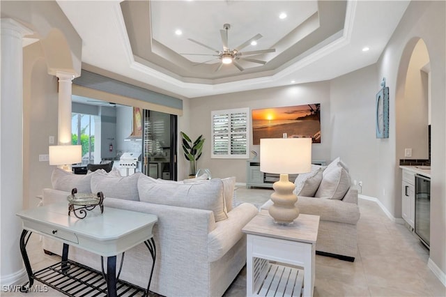 living room with ceiling fan, beverage cooler, a tray ceiling, and decorative columns