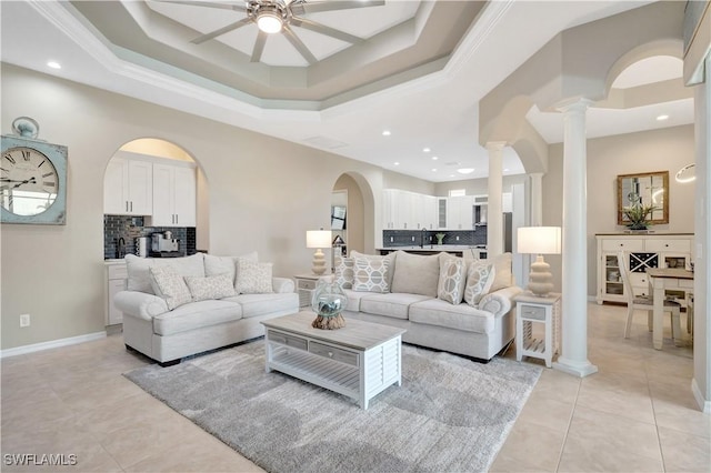 tiled living room featuring ceiling fan, sink, a tray ceiling, ornamental molding, and decorative columns