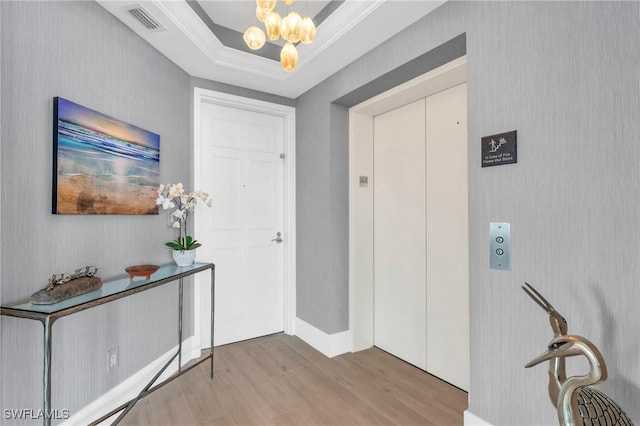 entryway featuring elevator, a raised ceiling, a notable chandelier, light wood-type flooring, and crown molding