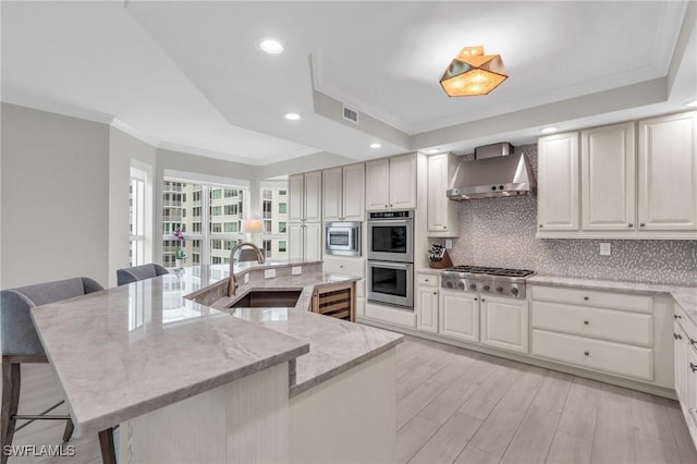 kitchen with wall chimney range hood, crown molding, appliances with stainless steel finishes, a sink, and a breakfast bar area