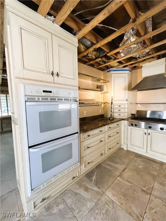 kitchen with cream cabinetry, white double oven, wall chimney exhaust hood, and stainless steel gas stovetop