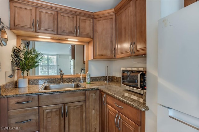 kitchen with a sink, dark stone counters, brown cabinetry, and freestanding refrigerator