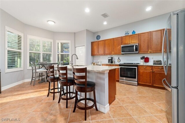 kitchen featuring appliances with stainless steel finishes, a breakfast bar, backsplash, light stone countertops, and a center island with sink