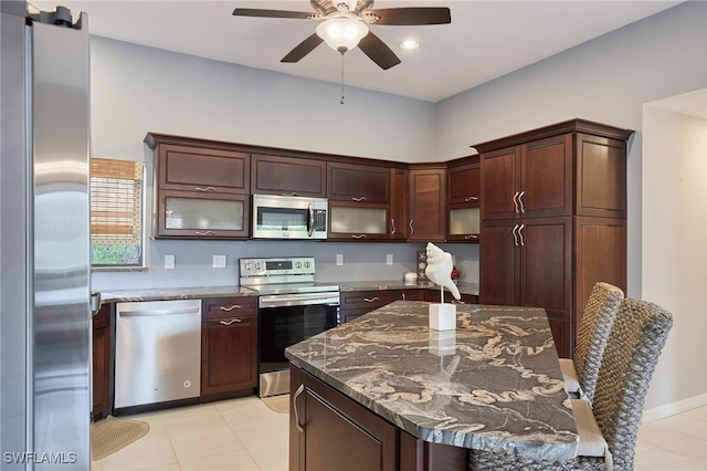 kitchen featuring light tile patterned floors, ceiling fan, appliances with stainless steel finishes, dark stone countertops, and dark brown cabinetry