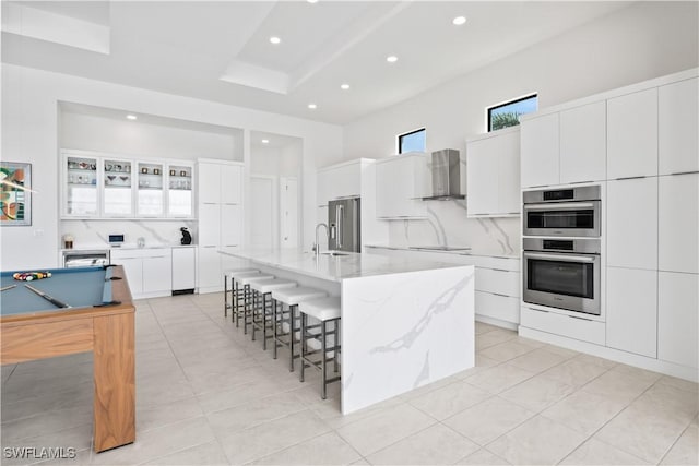 kitchen with white cabinetry, wall chimney exhaust hood, a large island with sink, and appliances with stainless steel finishes