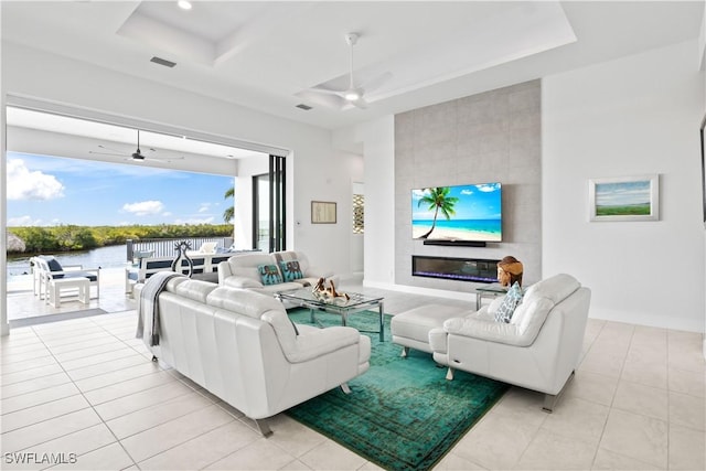 tiled living room with a water view, a large fireplace, ceiling fan, and a tray ceiling