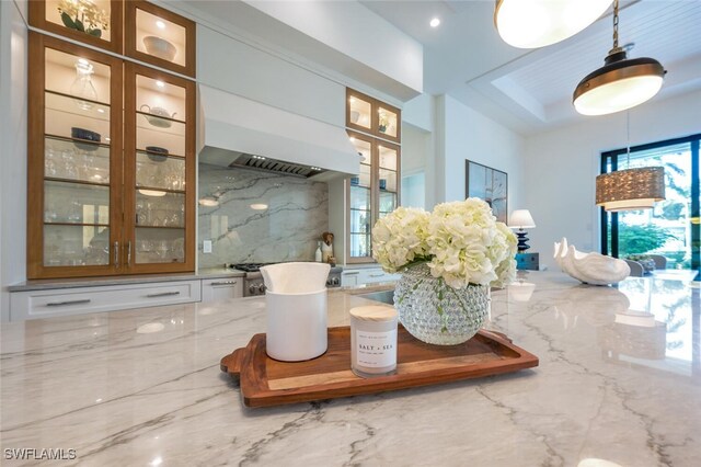 interior details with white cabinets, decorative backsplash, hanging light fixtures, light stone countertops, and custom range hood