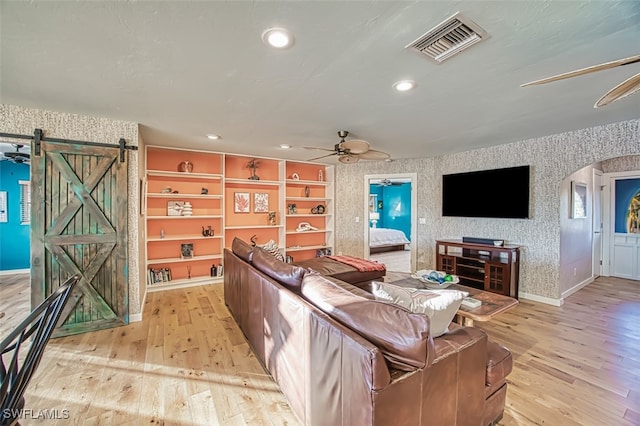 living room with built in shelves, light wood-type flooring, ceiling fan, and a barn door