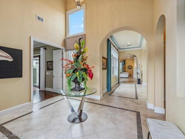 corridor with crown molding, light tile patterned floors, a towering ceiling, and a raised ceiling