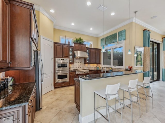 kitchen with appliances with stainless steel finishes, pendant lighting, dark stone countertops, ornamental molding, and a kitchen island with sink