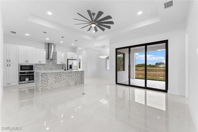 kitchen with appliances with stainless steel finishes, wall chimney exhaust hood, white cabinetry, ceiling fan, and a tray ceiling