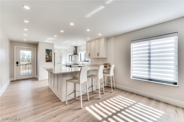 kitchen with white cabinets, wall chimney range hood, light stone counters, light hardwood / wood-style flooring, and a breakfast bar area