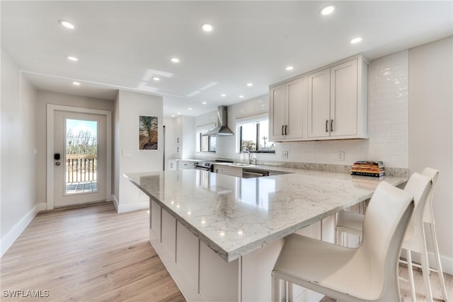 kitchen with light stone countertops, white cabinetry, wall chimney range hood, light wood-type flooring, and a breakfast bar